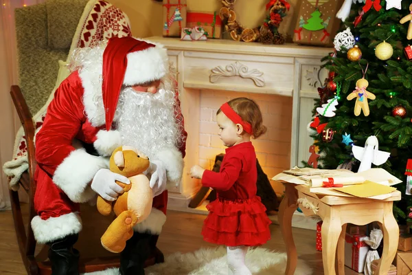 Санта-Клаус делает подарок маленькой симпатичной девочке у камина и елки дома — стоковое фото
