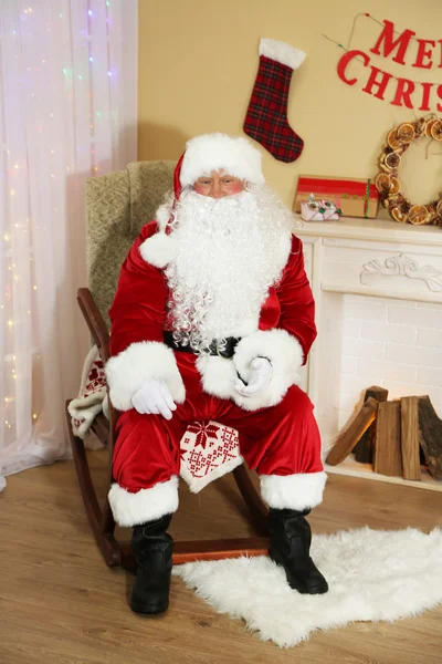 Santa claus zittend in een comfortabele stoel in de buurt van open haard thuis — Stockfoto