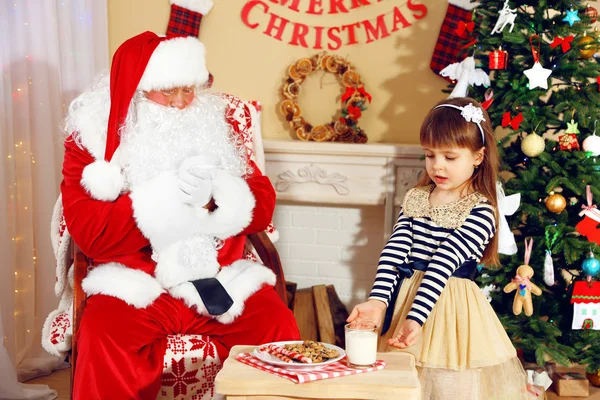 Little cute girl giving glass of milk to Santa Claus near Christmas tree at home — Stock Photo, Image