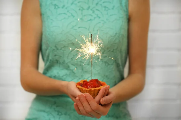 Woman holding tasty cake with sparkler, on grey wall background — Stock Photo, Image