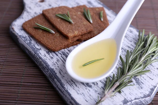 Crispbread with sprigs of rosemary on cutting board and spoon of oil on bamboo mat background