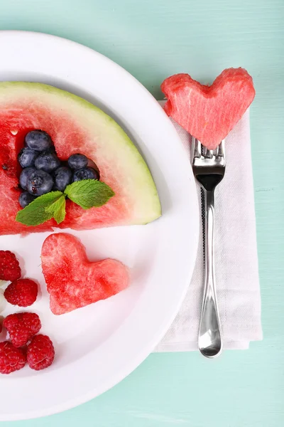 Frische saftige Wassermelonenscheibe mit ausgeschnittener Herzform, gefüllte frische Beeren, auf Teller, auf Holzgrund — Stockfoto