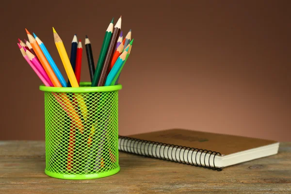 Colorful pencils in green metal holder with notebook on wooden table and shaded color background — Stock Photo, Image