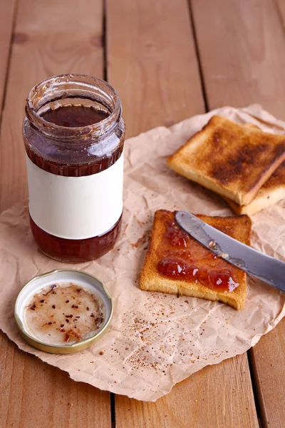 Kağıt parçasına reçel kavanoz ahşap masa arka plan üzerinde yakınındaki bıçakla yayılmış tost ekmeği — Stok fotoğraf