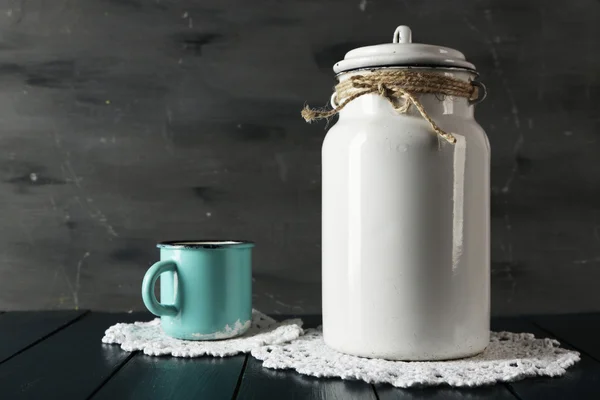 Milk can and cup on lace doily on wooden table and dark background — Stock Photo, Image