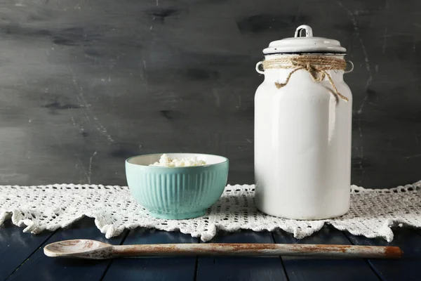 Milk can with bowl of cottage cheese on lace doily on wooden table and dark background — Stock Photo, Image
