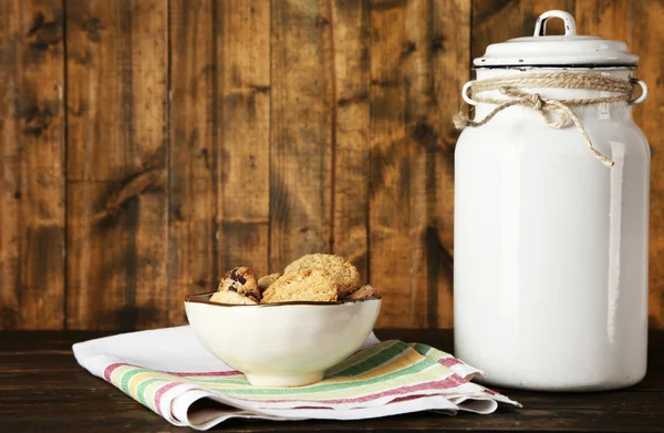 Milk can with bowl of cookies on dishcloth on rustic wooden background — Stock Photo, Image