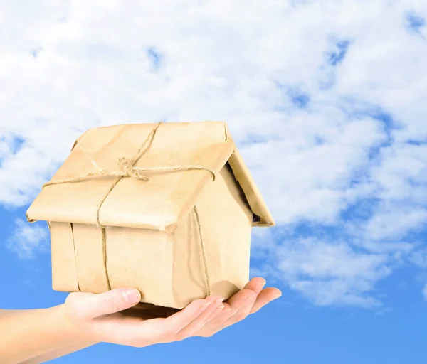 Manos de mujer sosteniendo la casa envuelta en papel kraft marrón sobre el fondo del cielo — Foto de Stock