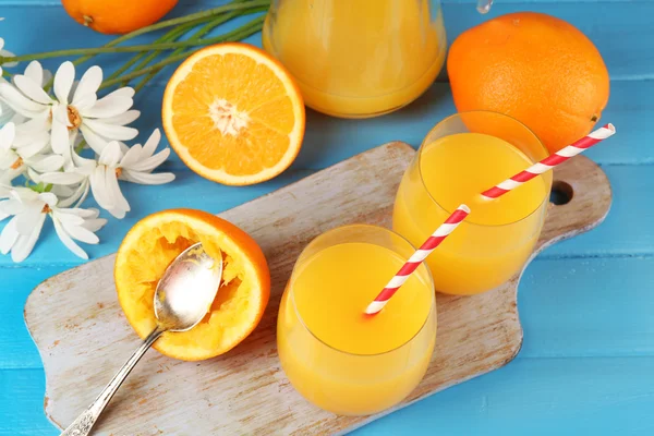 Bicchiere di succo d'arancia con fette, fiori e cannucce sul tagliere su sfondo di legno a colori — Foto Stock