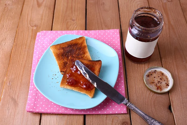 Pan tostado untado con mermelada en plato y servilleta con cuchillo cerca del frasco sobre fondo de mesa de madera — Foto de Stock