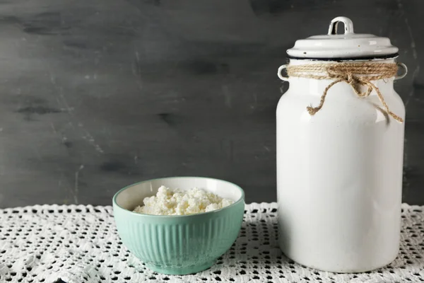 Milk can with bowl of cottage cheese on lace doily on wooden table and dark background — Stock Photo, Image