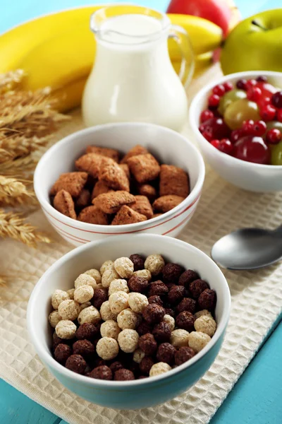 Diverses céréales sucrées dans des bols en céramique, fruits et cruche avec du lait sur la serviette, sur fond en bois de couleur — Photo
