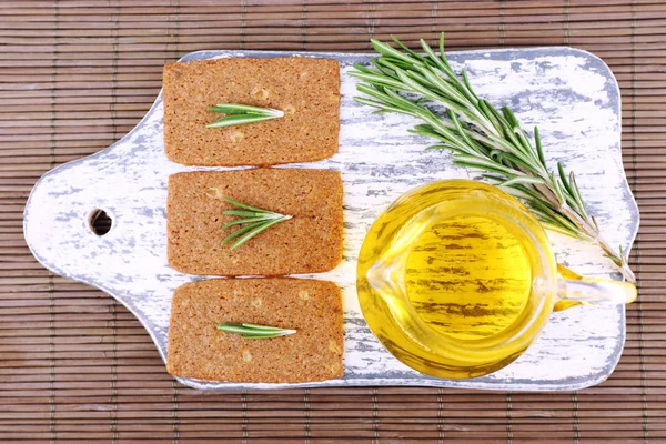 Crispbread with sprigs of rosemary on wooden cutting board with jug of oil on bamboo mat background — Stock Photo, Image