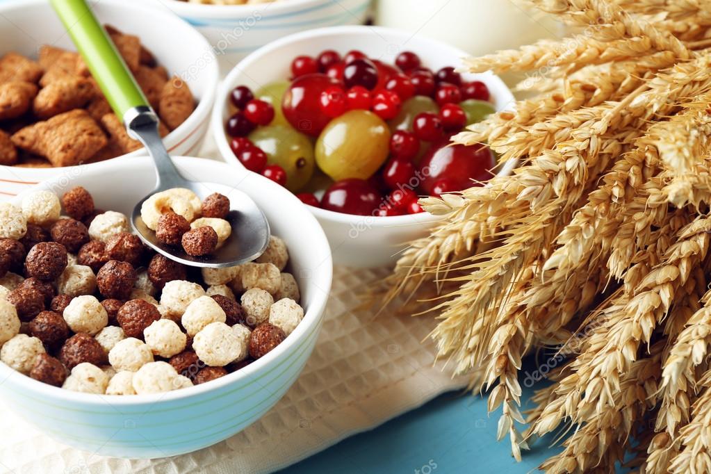 Various sweet cereals in ceramic bowls, fruits and jug with milk on napkin, on color wooden background