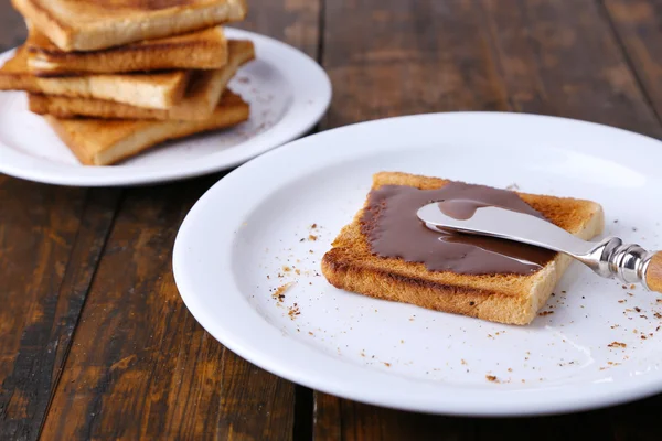 Pane tostato spalmato con cioccolato su piatto con coltello e tovagliolo su sfondo tavolo di legno — Foto Stock