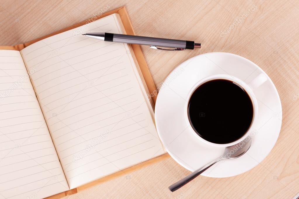 Cup of coffee on saucer with open notebook and pen on wooden table background