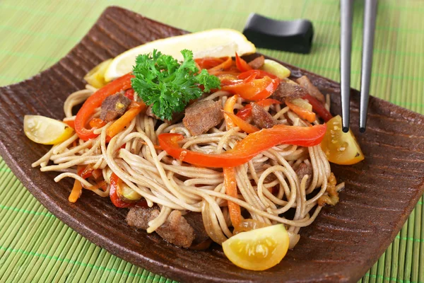 Macarrão chinês com legumes e carne assada na placa no fundo do tapete de bambu — Fotografia de Stock