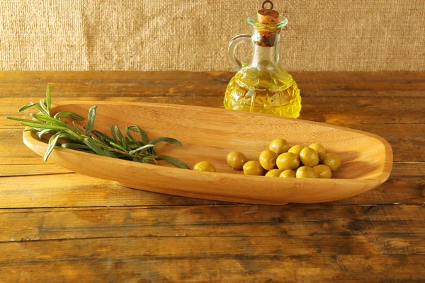 Green olives and branch in oblong bowl with oil can on rustic wooden table, on burlap background — Stock Photo, Image