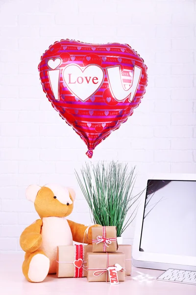 Urso de pelúcia com caixas atuais, planta e amor balão de coração na mesa de computador de madeira, no fundo da parede de tijolo — Fotografia de Stock