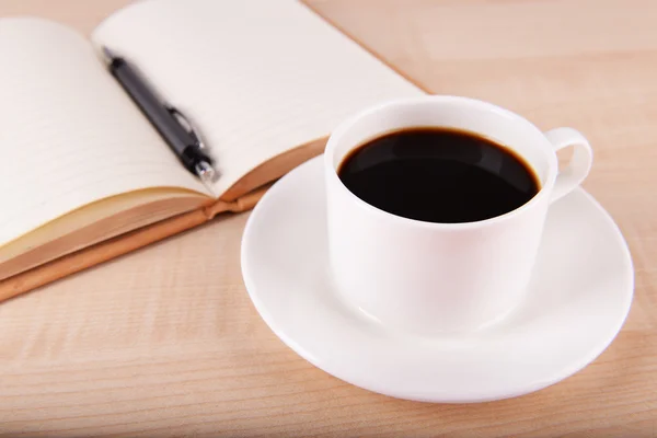 Tasse Kaffee auf Untertasse mit offenem Notizbuch und Stift auf Holztischhintergrund — Stockfoto