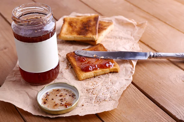 Toustového chleba potřete jam na kusu papíru s nožem u sklenice na dřevěný stůl pozadí — Stock fotografie