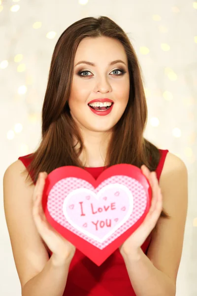 Chica sonriente sosteniendo la tarjeta de San Valentín con saludos en el fondo de luces —  Fotos de Stock