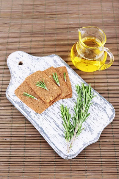 Crispbread with sprigs of rosemary on wooden cutting board with jug of oil on bamboo mat background — Stock Photo, Image