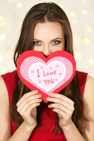 Chica sonriente sosteniendo la tarjeta de San Valentín con saludos en el fondo de luces — Foto de Stock