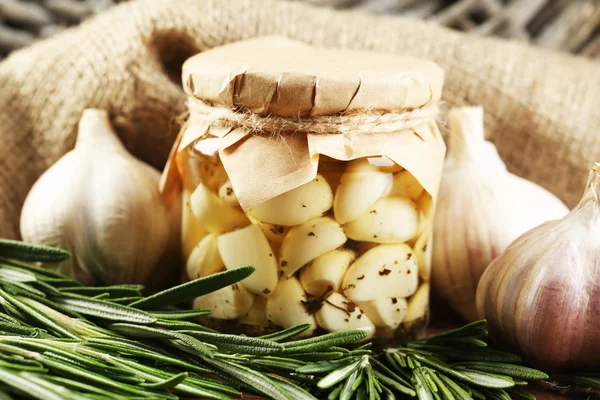 Canned garlic in glass jar and wicker mat and rosemary branches, on wooden background — Stock Photo, Image