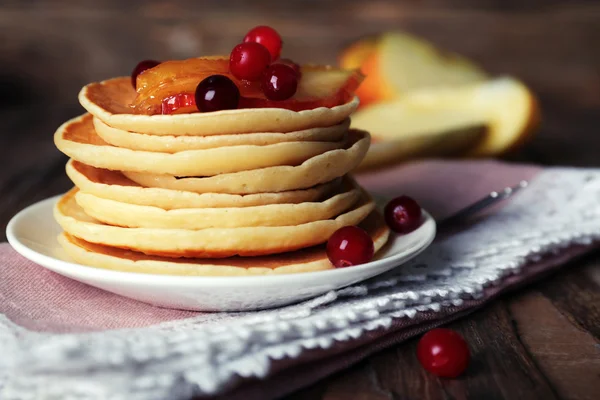 Стопка вкусных блинчиков с ломтиками яблока и ягод на тарелке и салфеткой на деревянном фоне — стоковое фото