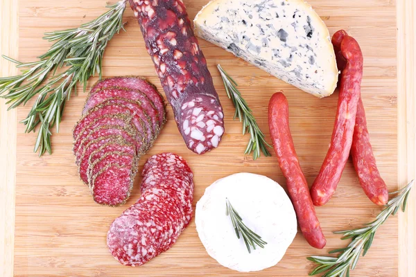 Uzené klobásy se sýrem, olivami a snítky rozmarýnu na dřevěné krájecí deska pozadí — Stock fotografie