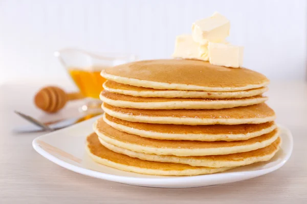 Pilha de deliciosas panquecas com manteiga e mel em pires na mesa e fundo leve — Fotografia de Stock