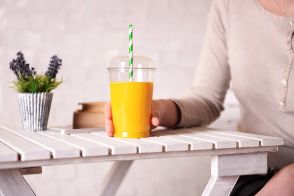 Female hand at wooden table with fast food closed cup of orange juice and near books and plant on light wall background