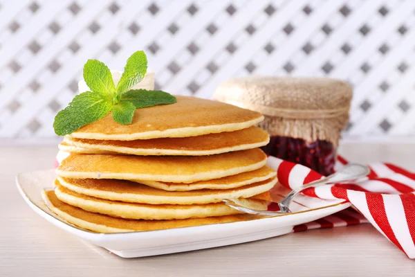 Pila di frittelle su piatto con banca di marmellata su tavolo e sfondo di reticolo di legno — Foto Stock