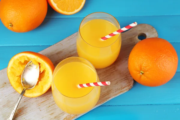 Kieliszek soku pomarańczowego z słomek, łyżka i plastry na deska do krojenia na kolor drewniane tła — Zdjęcie stockowe