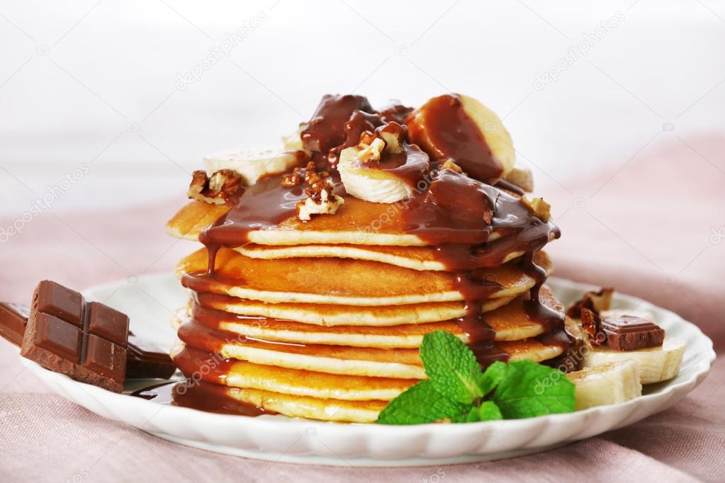 Stack of pancakes with mint, walnuts, chocolate and slices of banana on table with fabric on wooden planks background