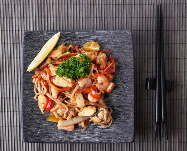 Fideos chinos con verduras y mariscos en plato sobre fondo de estera de bambú — Foto de Stock