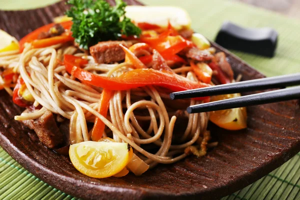 Chinese noodles with vegetables and roasted meat on plate on bamboo mat background — Stock Photo, Image
