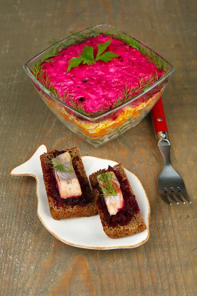 Ensalada de arenque ruso en tazón de vidrio y sándwiches con arenque salado, sobre fondo de mesa de madera — Foto de Stock