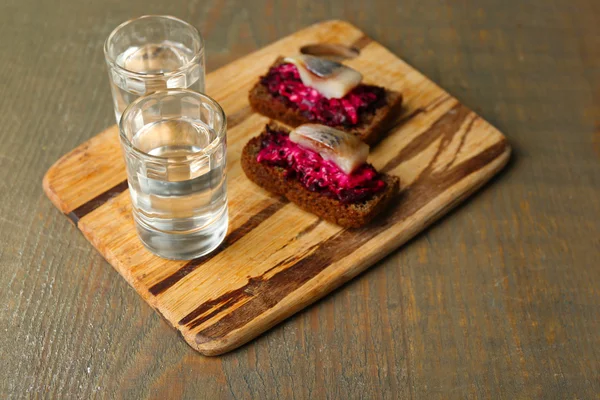 Canape herring with beets on rye toasts, on wooden board, and glass of vodka on wooden table background — Stock Photo, Image