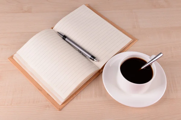 ノートブックを開くとソーサーにコーヒーと木製のテーブル背景にペン — ストック写真