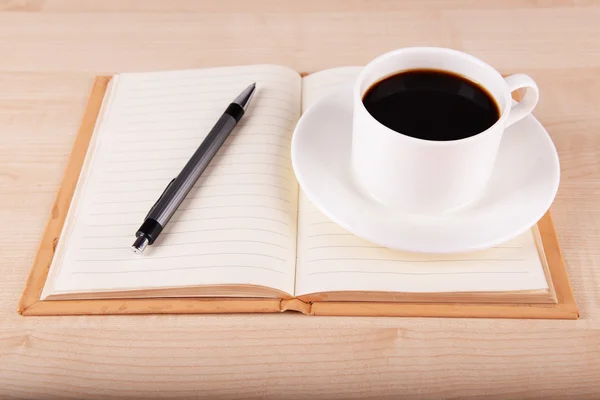 Tazza di caffè su piattino con notebook aperto e penna su sfondo tavolo in legno — Foto Stock