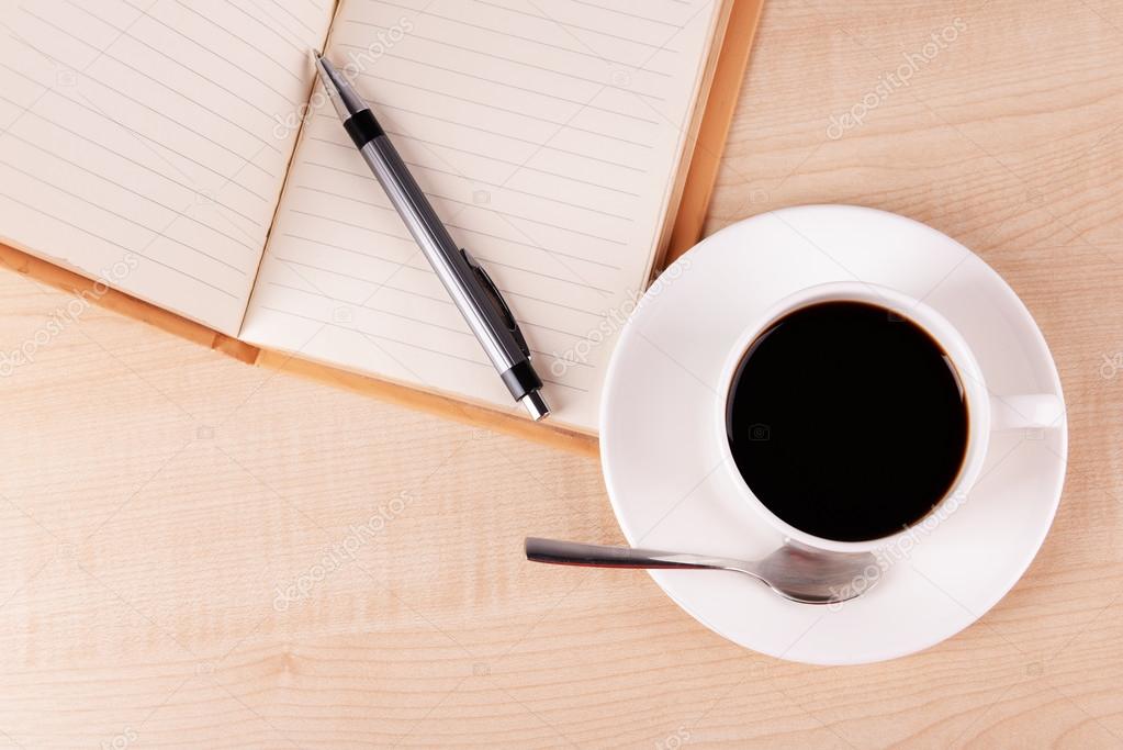 Cup of coffee on saucer with open notebook and pen on wooden table background