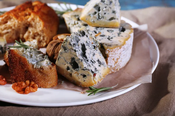 Blue cheese with sprigs of rosemary, bread and nuts on plate with burlap cloth and color wooden table background — Stock Photo, Image