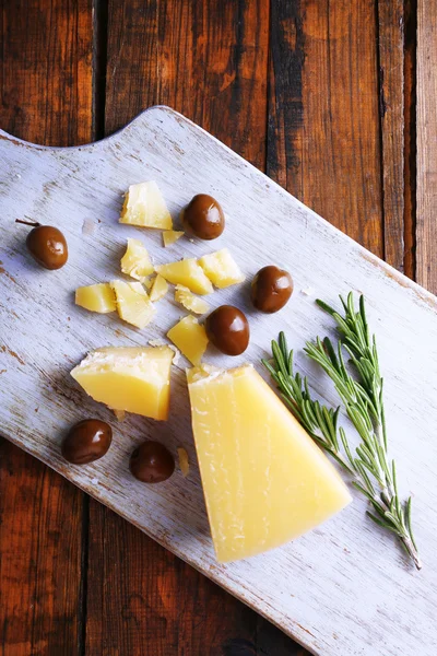 Parmesan cheese on cutting board with sprig of rosemary and olives on wooden table background — Stock Photo, Image