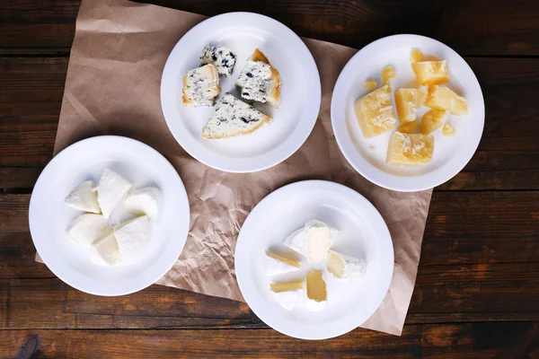 Plates of various types of cheese on sheet of paper on wooden table background — Stock Photo, Image