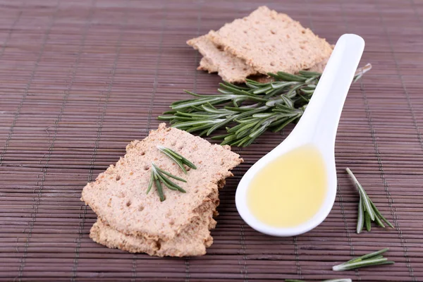 Crispbread with sprigs of rosemary and spoon of oil on bamboo mat background — Stock Photo, Image