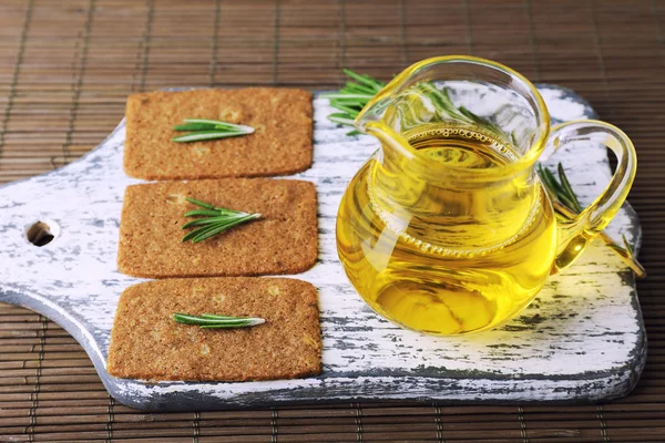 Crispbread com raminhos de alecrim em tábua de corte de madeira com jarro de óleo sobre fundo de tapete de bambu — Fotografia de Stock