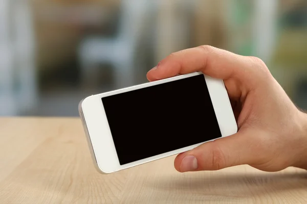 Mano que sostiene el teléfono móvil inteligente en la mesa de madera y el fondo borroso claro —  Fotos de Stock