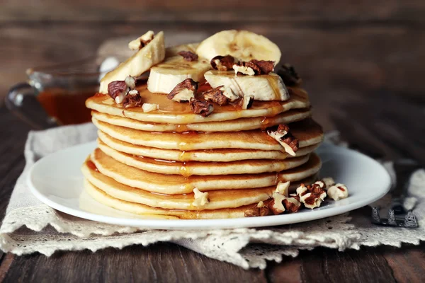 Pilha de deliciosas panquecas com chocolate, mel, nozes e fatias de banana no prato e guardanapo no fundo de madeira — Fotografia de Stock